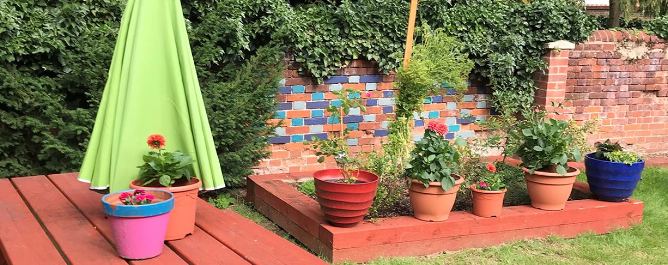 Vibrant garden area, with painted brickwork behind a red painted risen planter, matching bench and potted plants.