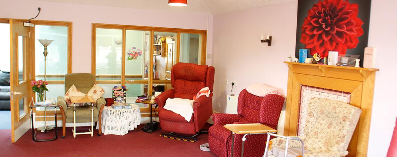 A broad seating area, with glass panelled wall looking out into the corridor, Featuring a mixed bunch of seating options, from a red leather electric armchair, red and white patterned reclining chair, and sturdy traditional patterned armchairs.