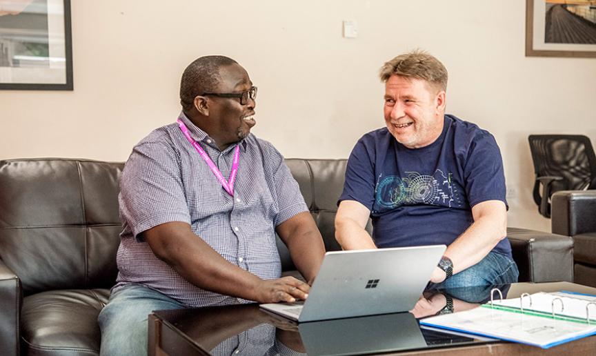 A supported service user and a member of staff sat together and smiling, they are sat on a brown leather sofa, in front of a laptop on top of a coffee table.