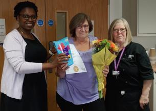 Sanctuary chef pictured in a purple top, holding a certificate, a box of Roses chocolates and a bouquet of flowers. She stands between two colleagues in an indoor setting.