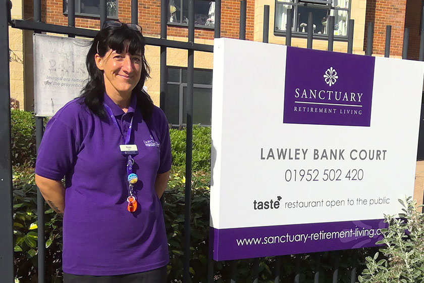 Wellbeing and Inclusion Assistant stands in front of the sign for Lawley Bank Court, in a purple Sanctuary uniform top