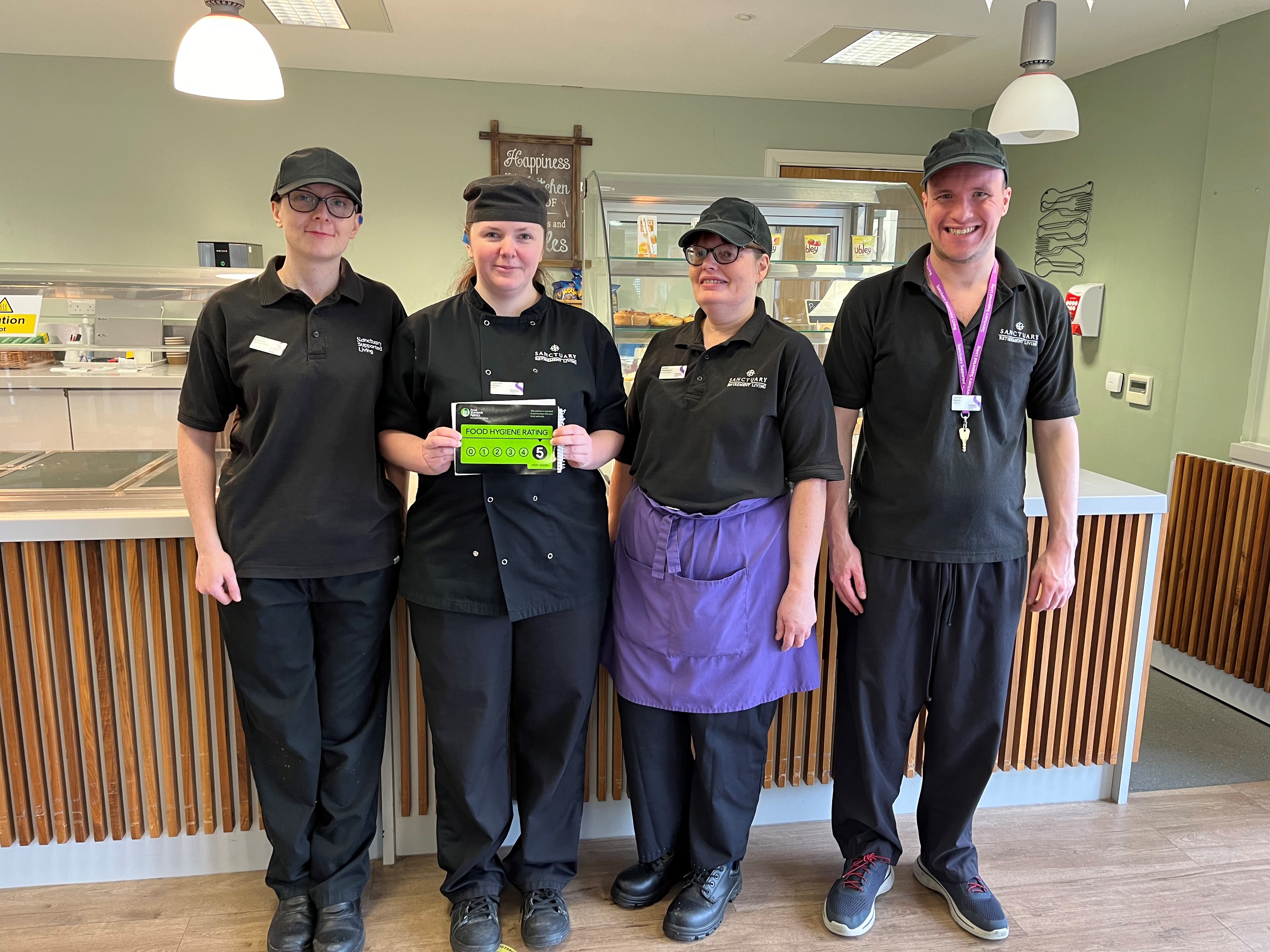 Baskeyfield House catering team celebrate Four people wearing black catering uniform, standing holding a 5 rating from the Food Standards Agency and smiling at the camera.
