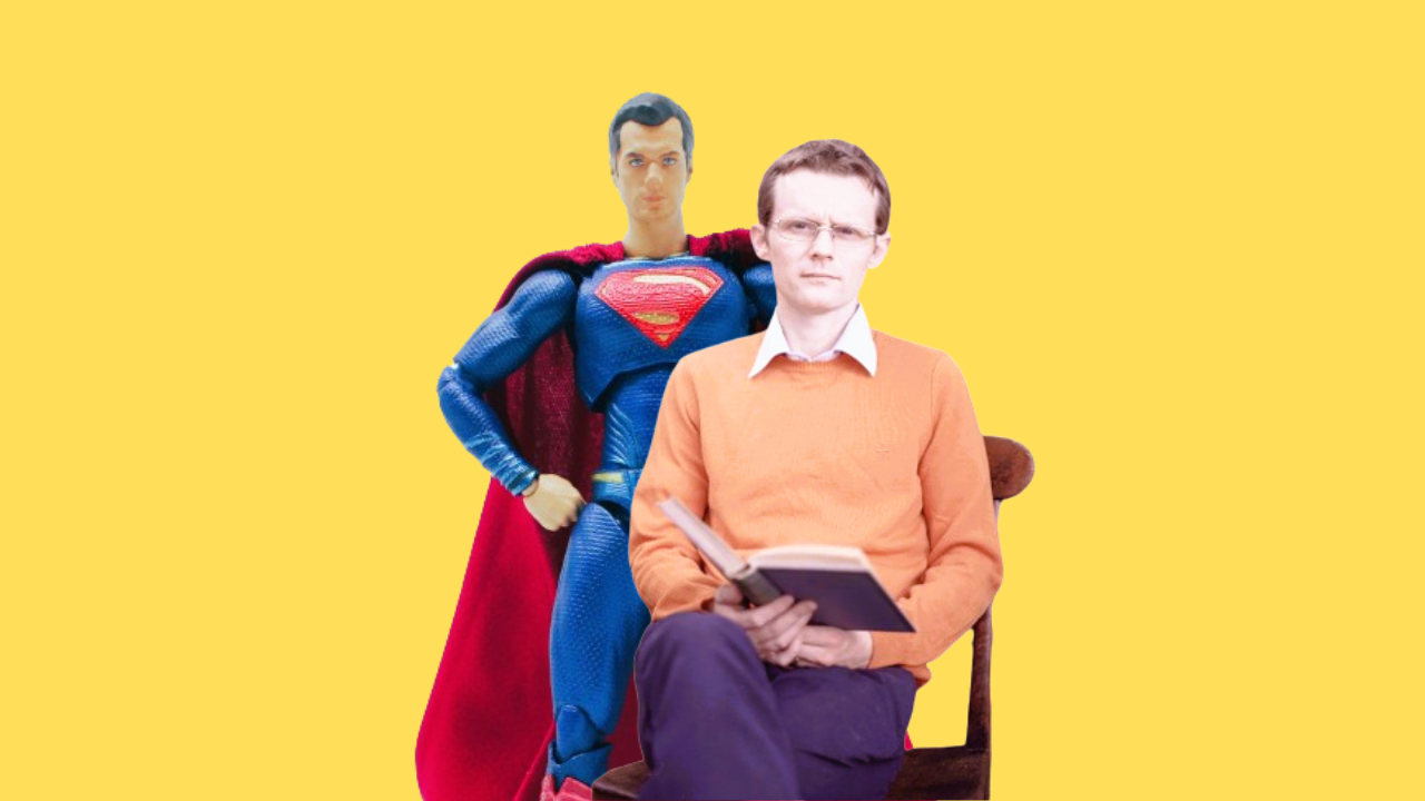 An image of Neil, a man sat wearing an orange jumper holding a book in front of a Superman figure.