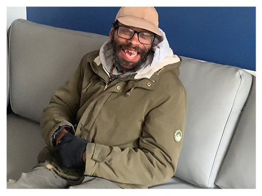 Andy A black man wearing a green coat and hat, sat on a grey sofa against a blue wall, smiling at the camera