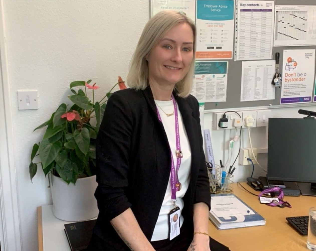 A white woman with blonde hair, wearing a dark suit jacket and white top, with a pink lanyard round her neck, sat at a desk in an office and smiling at the camera.