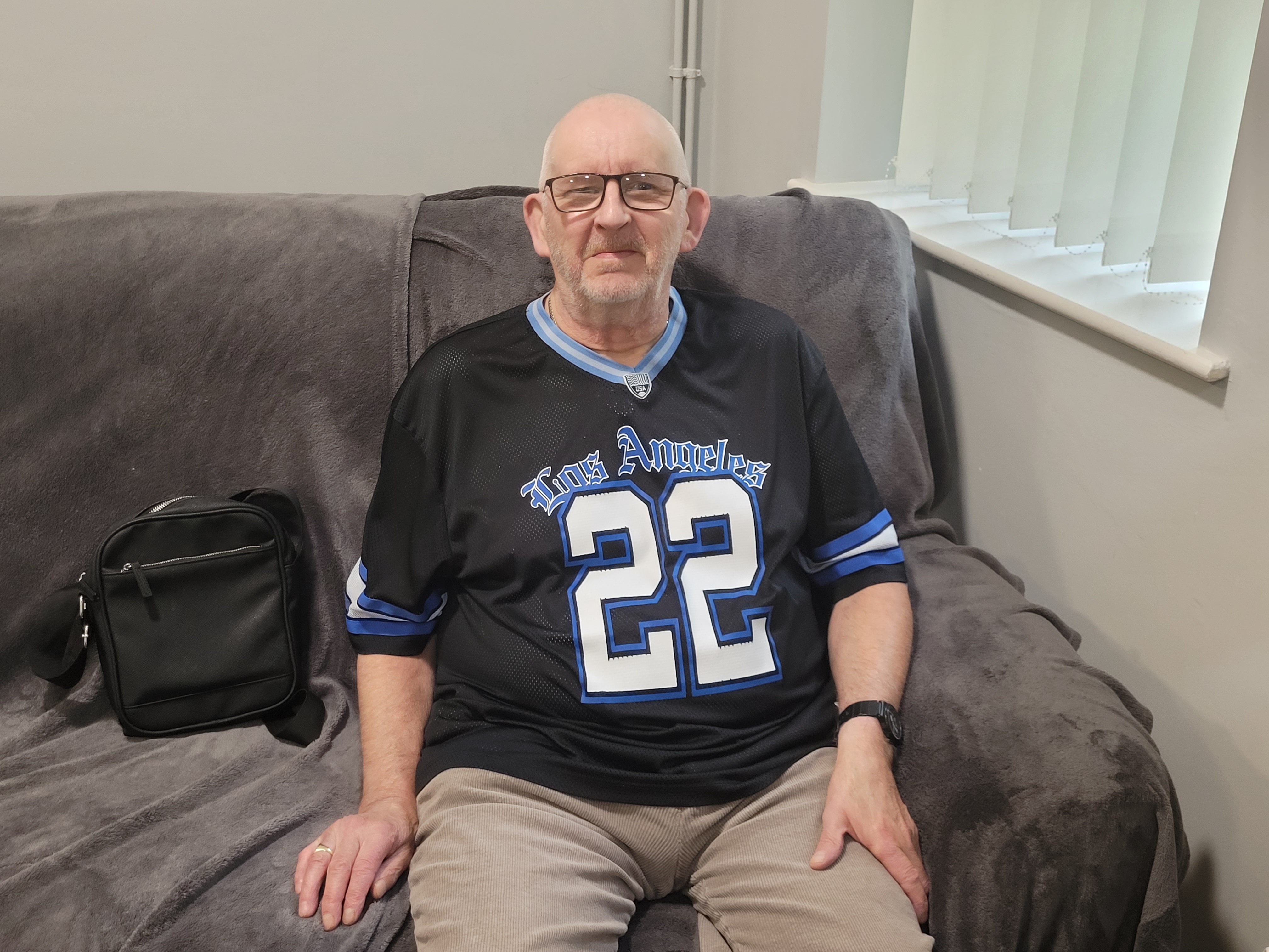 An older white man wearing glasses and a dark Los Angeles sports shirt, sitting on a cream sofa and smiling at the camera.