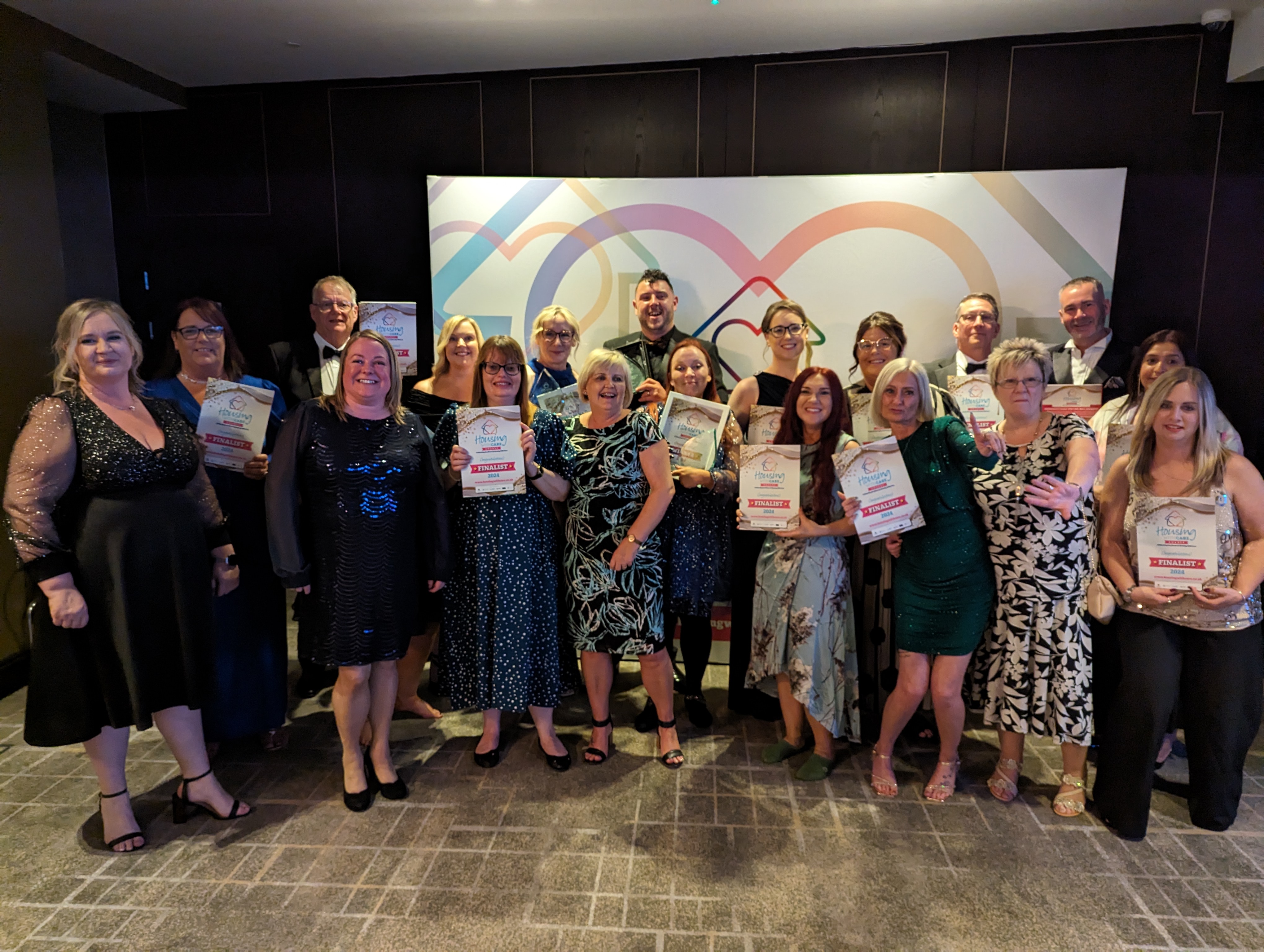 Quadruple award win for Sanctuary Supported Living A group of 19 people standing together and smiling at the camera, wearing party outfits and suits, holding up certificates and awards.