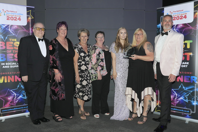 A group of seven individuals from Sanctuary Supported Living who appear to be of white ethnicity, consisting of five women and two men, are dressed in formal attire and standing in front of two ‘Winners’ pull-up banners. They are all smiling and one person holds an octagonal, glass Supported Housing award.