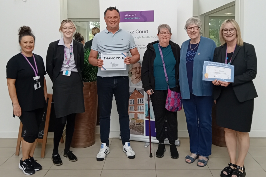 Five women standing in a row with one man in the centre, smiling at the camera and holding a cheque.