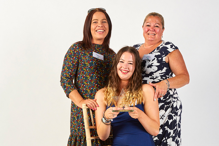 Three white women in evening dresses, two standing and one sitting in front, smiling for the camera and holding a glass trophy award.