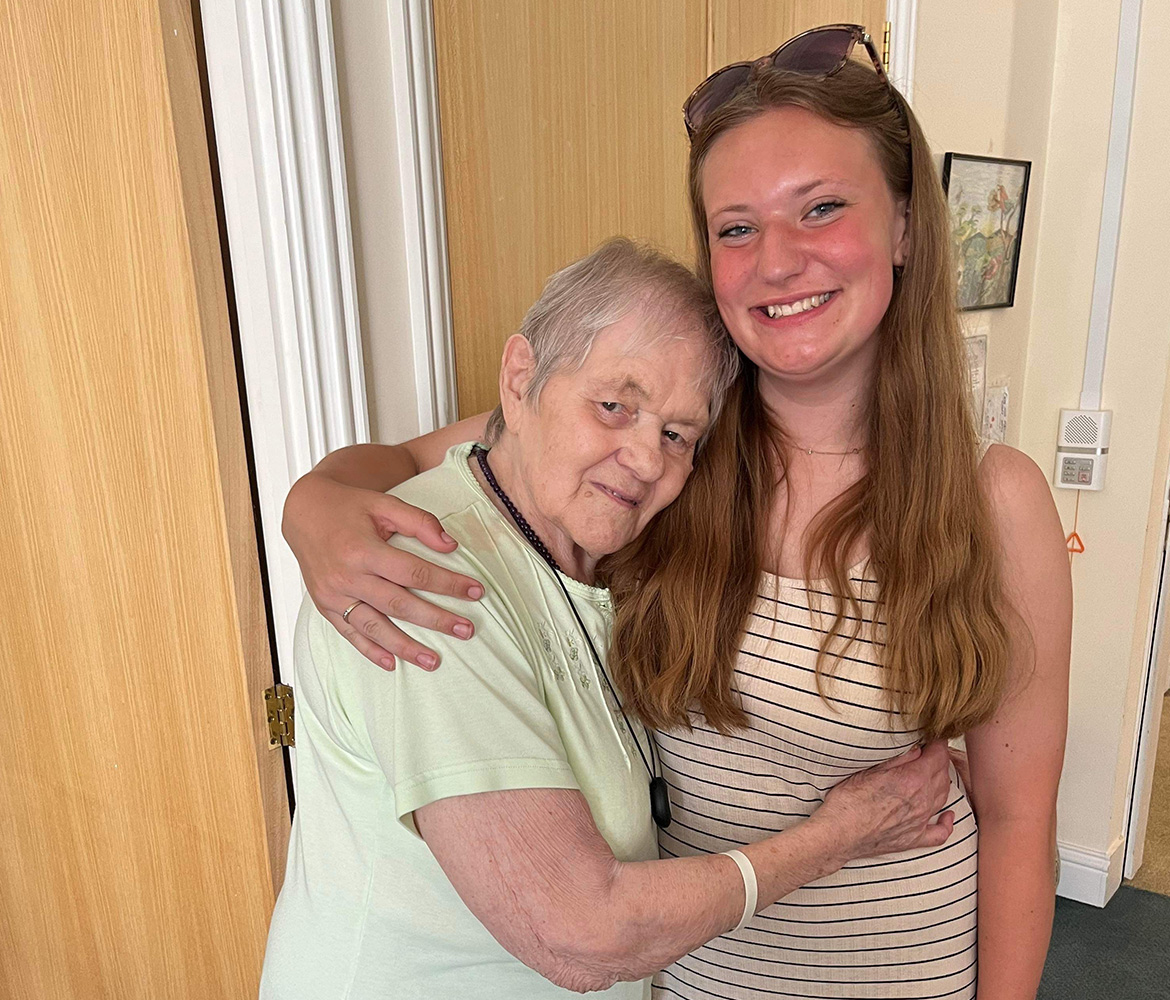 An older woman with short hair and a light green top hugging a young woman with long brown hair wearing a striped dress. They are standing indoors, smiling, with a wooden door and a painting in the background