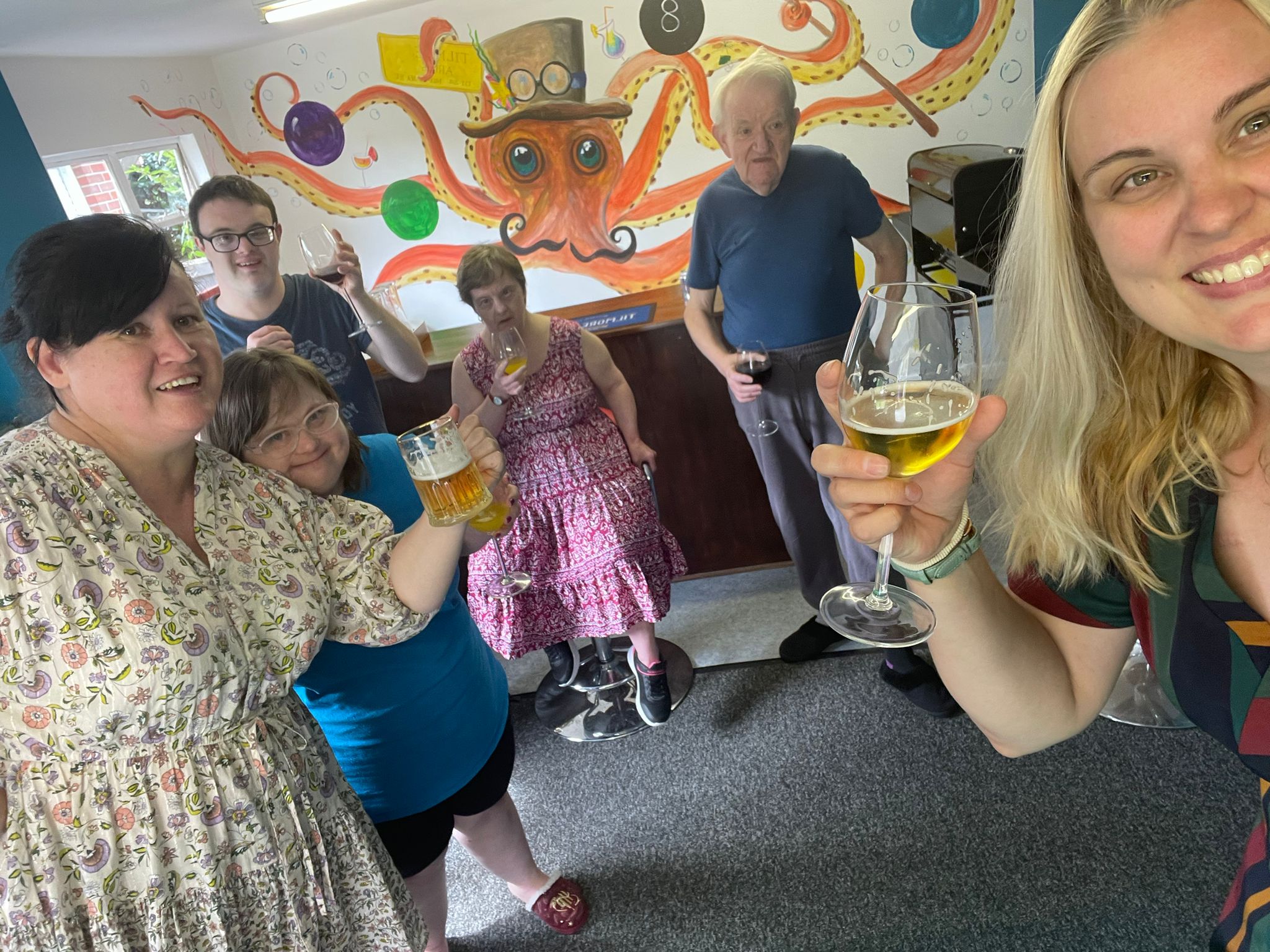 A group of six white men and women in their 30s, 40s and 50s, standing in a room decorated like a pub, each holding a drink and smiling to the camera.