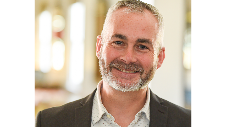 Headshot of a man wearing a shirt with a pattern of small dots and a dark suit jacket, smiling at the camera. The background is an indoor space with soft lighting, suggesting a professional setting