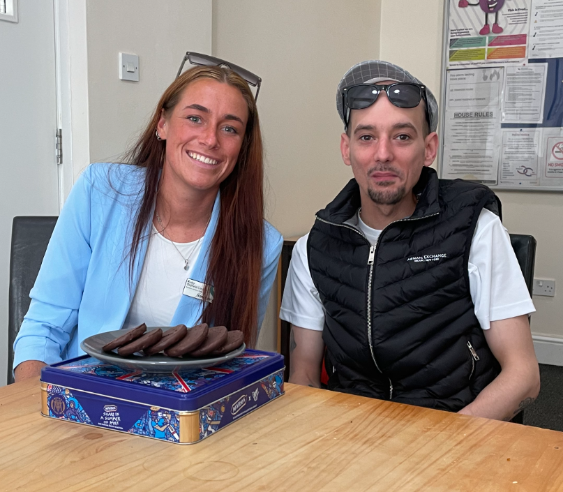 A young woman wearing a sky blue blazer with long auburn hair smiles alongside a young man with a goatee-style moustache and beard, wearing a black gilet and cap. Both have sunglasses resting on their heads, and there is a plate of chocolate biscuits in front of them.