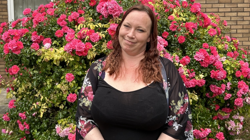 Young woman with mid-length hair in front of a bush full of pink flowers, clasping her hands together and smiling for the camera.