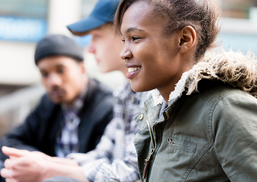 A young, black woman in a green jacket with a fur-lined hood sits outdoors smiling, while two out-of-focus young, white men in casual clothing talk in the background.