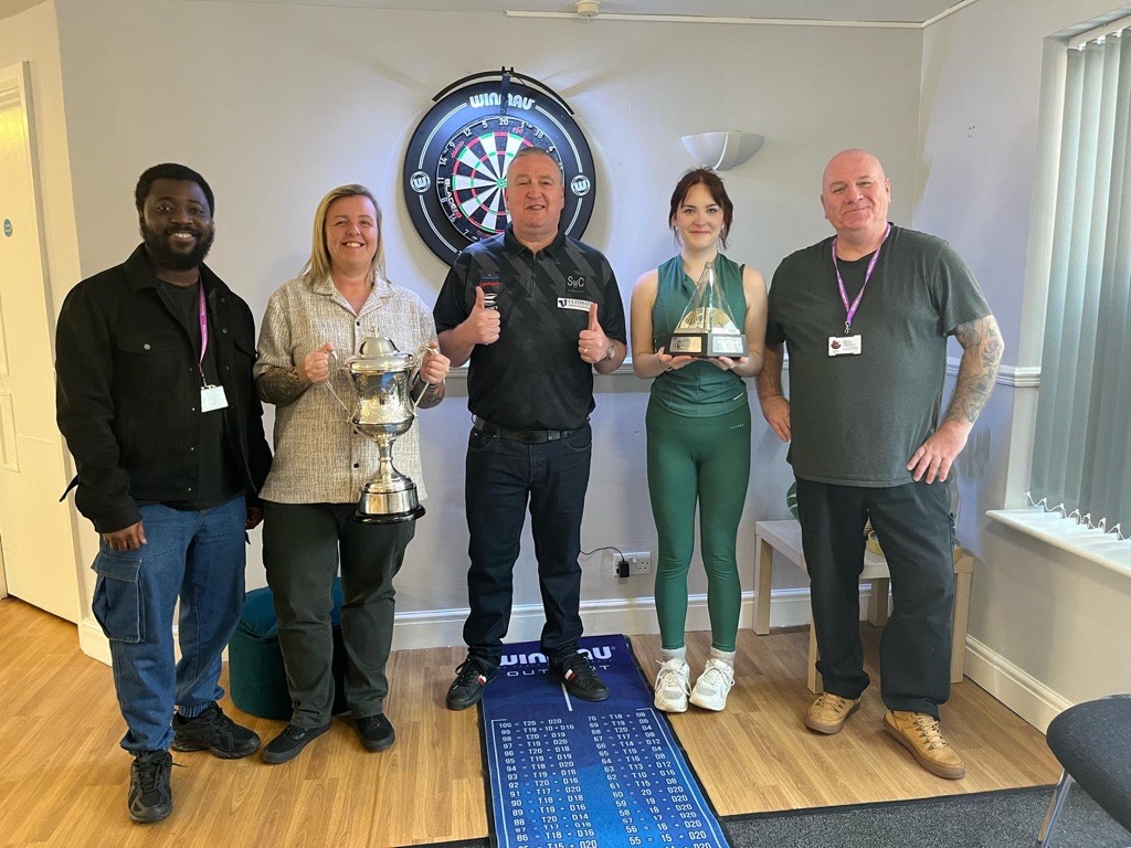 A group of five people stand together in a room with a dartboard mounted on the wall behind them.