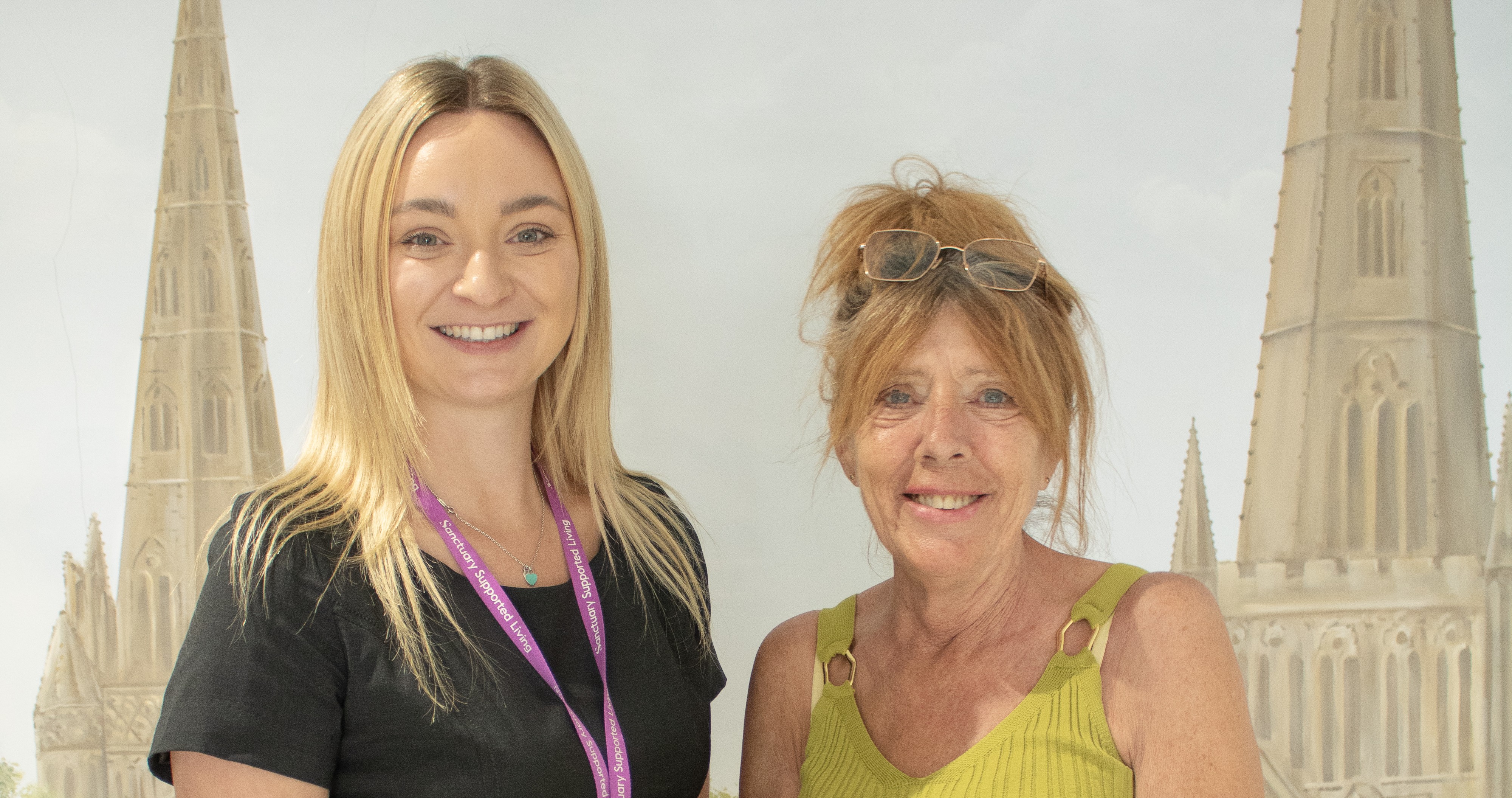 A young woman in a black dress with a lanyard stands next to an older woman wearing a green top and patterned skirt. The older woman is holding a bouquet of yellow roses. They are posing in front of a backdrop featuring tall, pointed church spires and a green landscape. Both women are smiling.