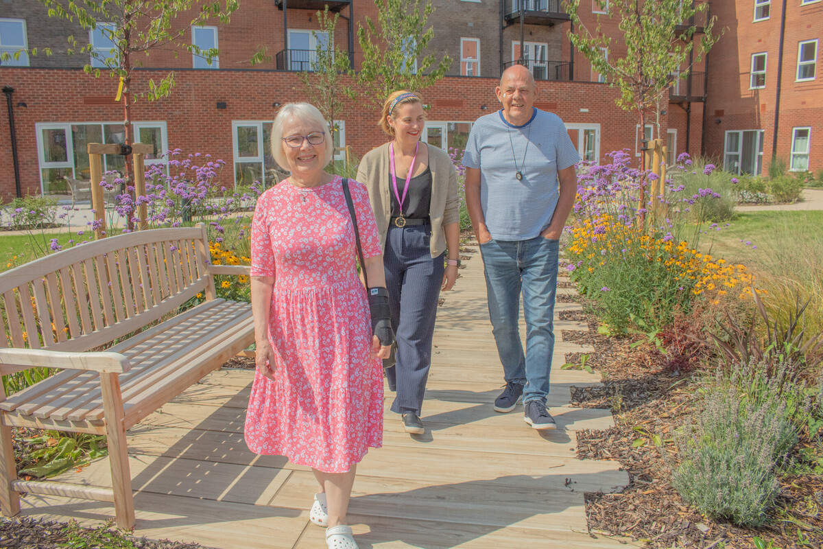 Three people walk in a sunny garden, on the left a happy older woman with glasses and short white hair wearing a pink floral dress. In the centre, a woman working for Sanctuary with brown hair, wearing a casual outfit. On the right, a cheerful man wearing a casual grey shirt and jeans. The garden is filled with colourful flowers and greenery, creating a vibrant atmosphere.