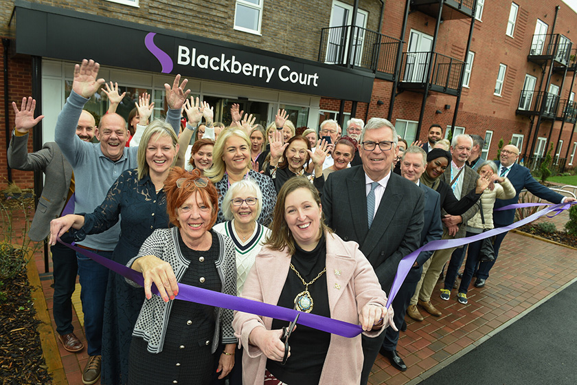A group of people gathered outside a modern brick building with balconies, standing in front of a sign that reads “Blackberry Court.” They are participating in a ribbon-cutting ceremony, holding a long purple ribbon stretched across the entrance. Many individuals have their hands raised in celebration, and the setting appears to be the opening of a new facility or building.