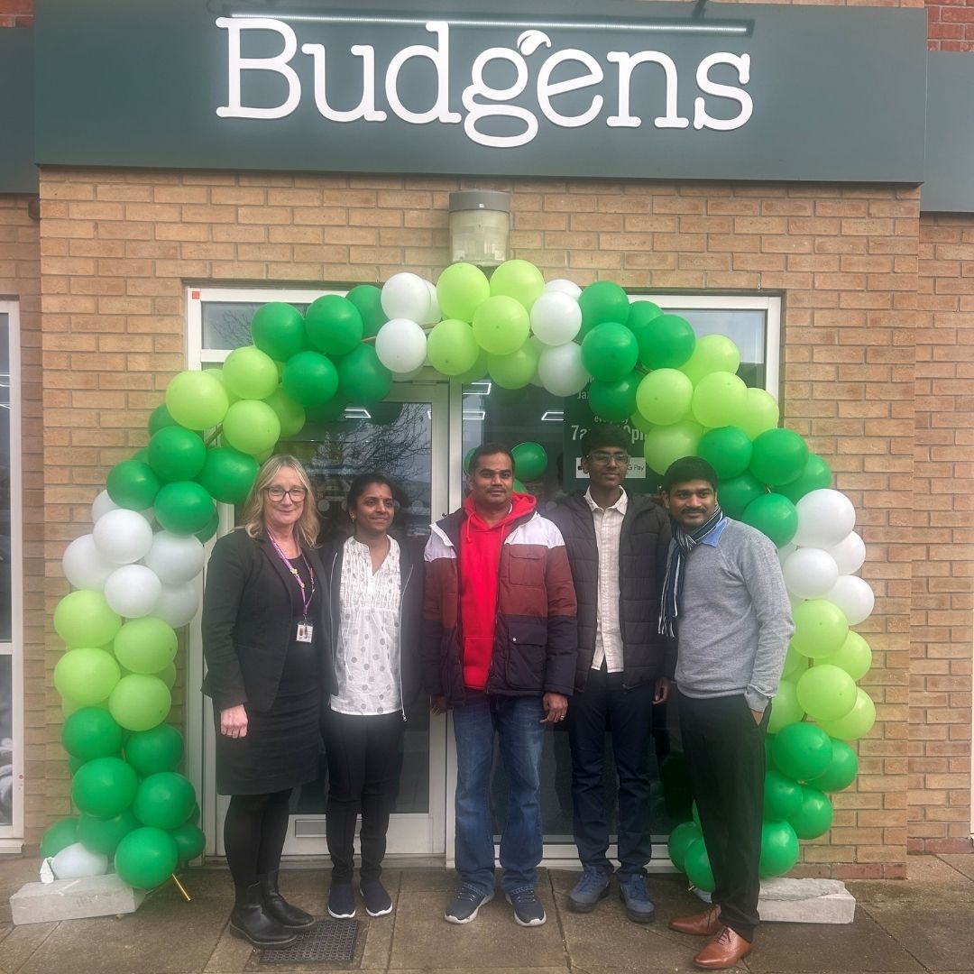 A group of five people stands in front of a Budgens store, which features a large arch made of green and white balloons. The individuals are smiling and dressed in casual clothing. The store's sign is prominently displayed above the entrance. The setting appears to be a festive occasion, likely a store opening or celebration.