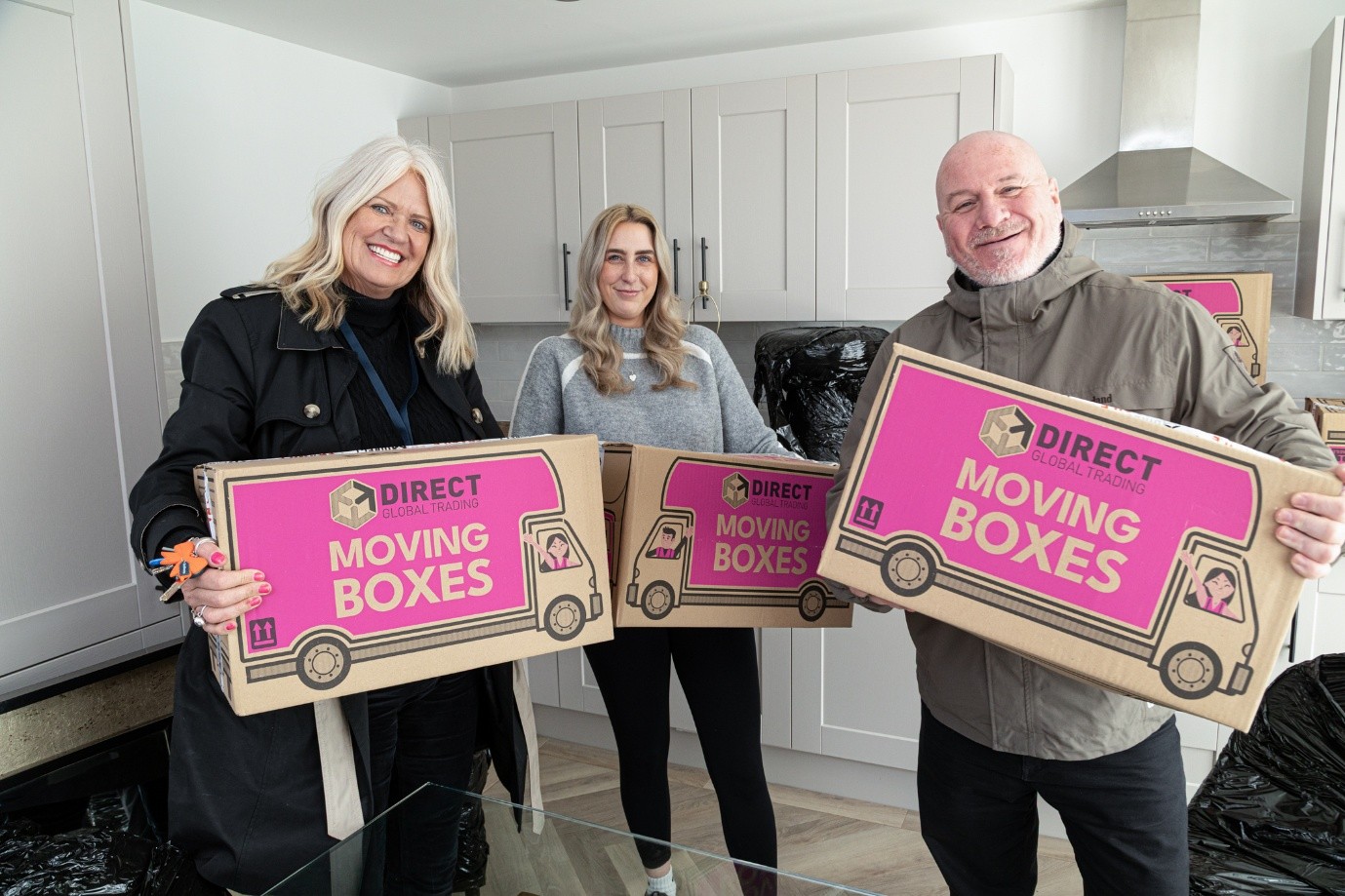 A group of three people smiling and standing in a modern kitchen, each holding a large cardboard box.