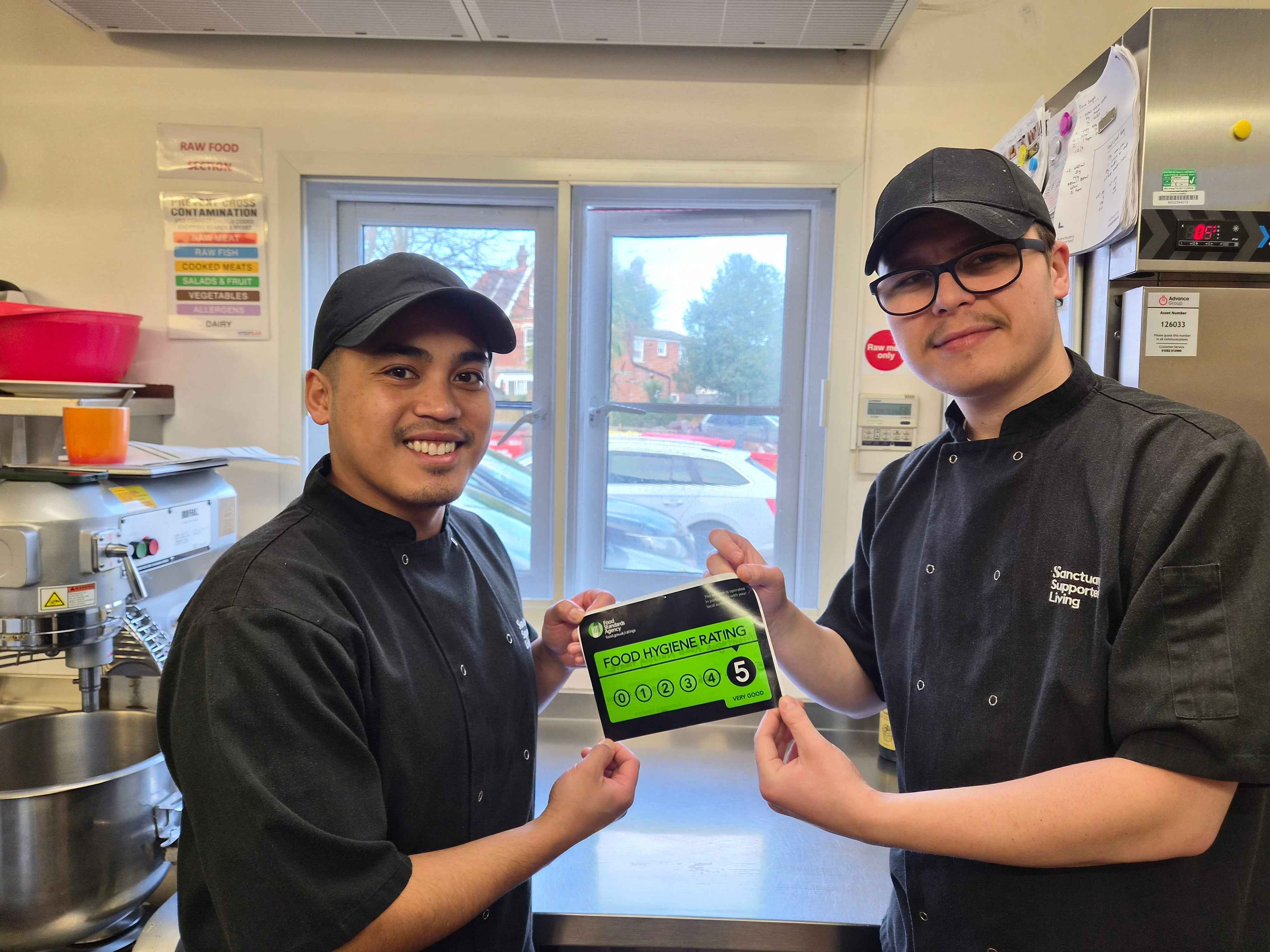 Two chefs in black uniforms and caps are standing in a kitchen. They are smiling and holding up a green food hygiene rating certificate that displays a score of 5. The kitchen is equipped with various cooking appliances and utensils, and there is a window in the background showing a view of parked cars outside.