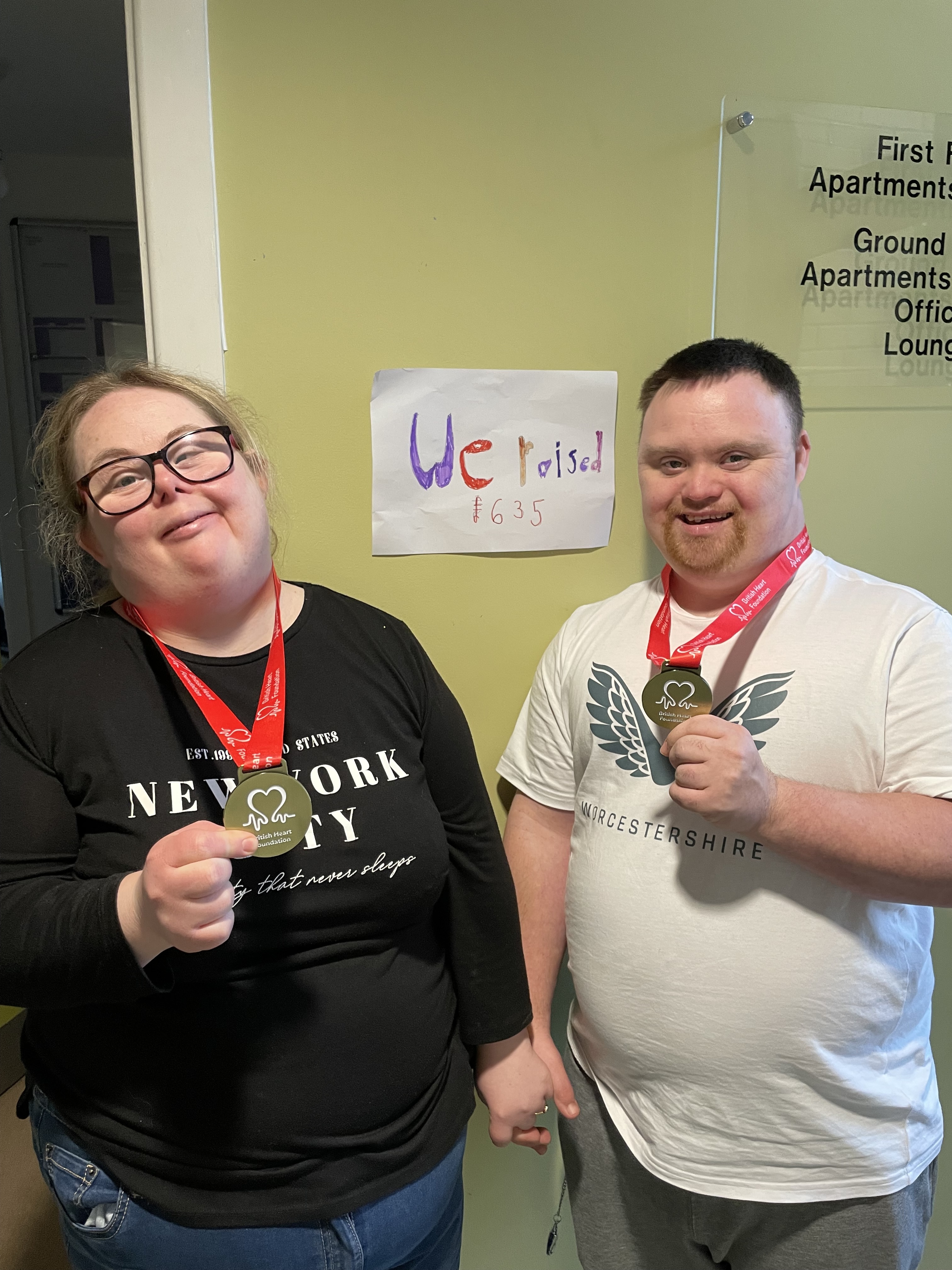 A woman and a man stand side by side, both smiling and holding medals. They are wearing red lanyards around their necks. Behind them is a sign that reads "We raised 1635," indicating a fundraising achievement.