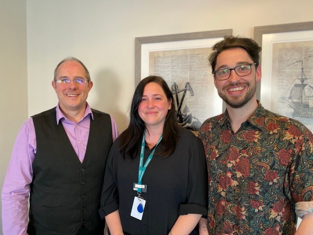 Three members of the Sanctuary Supported Living Learning and Development team standing indoors. One person wears a lilac shirt with a dark waistcoat, one wears a dark long‑sleeved top with a green lanyard, and one wears a patterned short‑sleeved shirt. They are standing side by side in front of framed artwork on a light‑coloured wall.