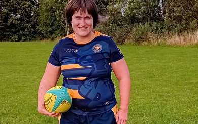 A woman wearing a rugby kit, holding a rugby ball and smiling at the camera.
