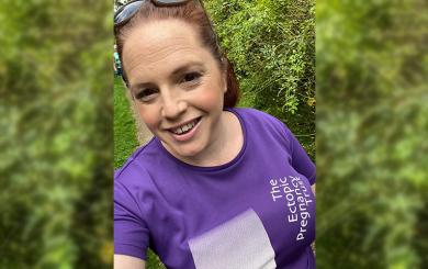 A woman wearing a purple tshirt, walking and smiling while taking a selfie.