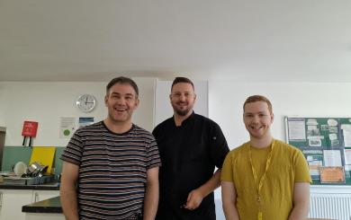 Three men stood in a kitchen and smiling at the camera, one wearing a chef's uniform.