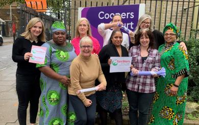 A group of nine women standing together in the street by a large sign that reads 'Coopers Court', smiling at the camera and holding signs that read 'FareShare'.