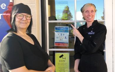Two women smiling at the camera, pointing to a sign on a door that shows a rating of 5 from the Food Standards Agency.