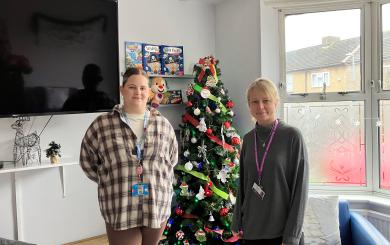 Two individuals standing next to a decorated Christmas tree in an indoor setting with children's books mounted on the wall behind them and a window letting in natural light
