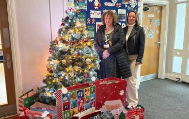 Two women stood by a Christmas tree and smiling at the camera, surrounded by gift bags containing presents.