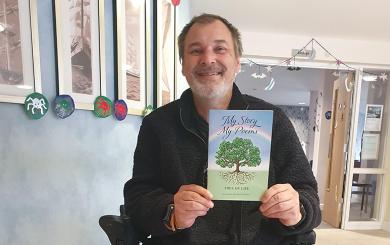 A bearded man proudly displays his poetry book titled "My Story, My Poems, Tree of Life." There is a rainbow on the front cover with a tree underneath