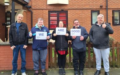 Five people standing in a row outside a building, smiling at the camera and holding signs saying 'We Are Good