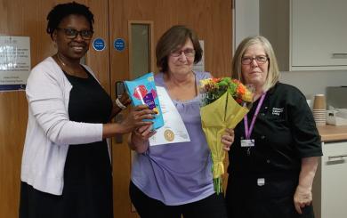 Amanda is in a purple top, holding a certificate, a box of Roses chocolates and a bouquet of flowers. She stands between two colleagues in an indoor setting