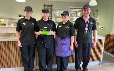 Four people wearing black catering uniform, standing holding a 5 rating from the Food Standards Agency and smiling at the camera.