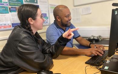 A woman and a man sat at a desk, looking at a computer together.