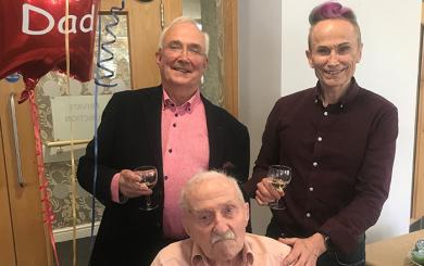 Three smartly dressed gentlemen pose for a photograph next to a red, star-shaped balloon that reads "Happy 100th Dad." The gentleman at the front is sitting in a wheelchair. He is elderly and wears a pastel pink shirt. The gentleman on the left behind him wears a salmon pink shirt and glasses and the gentleman on the right wears a purple mauve shirt, with a streak of purple in his hair to match. They both smile broadly with wine glasses in their hands.