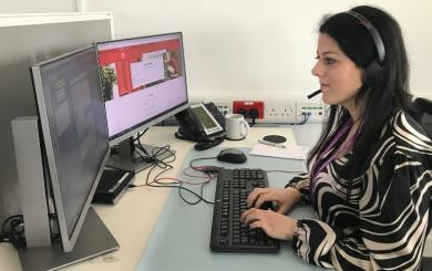A member of Sanctuary Supported Living staff sat at a desk working on a computer whilst wearing a telephonw headset