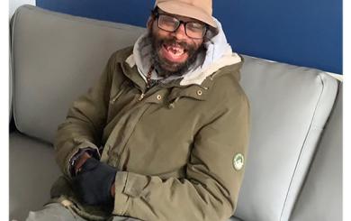 A black man wearing a green coat and hat, sat on a grey sofa against a blue wall, smiling at the camera