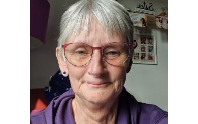 A white woman in her 70s, sat on a sofa in her flat, smiling at the camera.