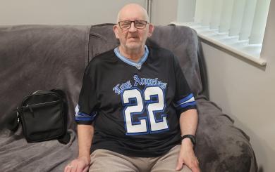  An older white man wearing glasses and a dark Los Angeles sports shirt, sitting on a cream sofa and smiling at the camera.