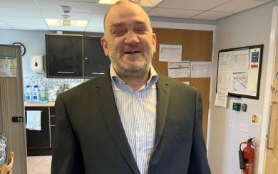 A middle-aged white man with shaved hair and stubble, wearing a dark grey suit and white striped shirt, looking at the camera 