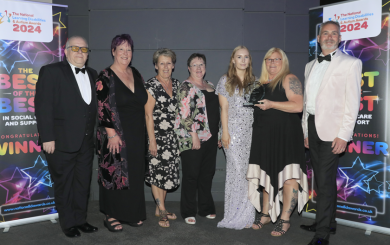 A group of seven individuals from Sanctuary Supported Living who appear to be of white ethnicity, consisting of five women and two men, are dressed in formal attire and standing in front of two ‘Winners’ pull-up banners. They are all smiling and one person holds an octagonal, glass Supported Housing award.