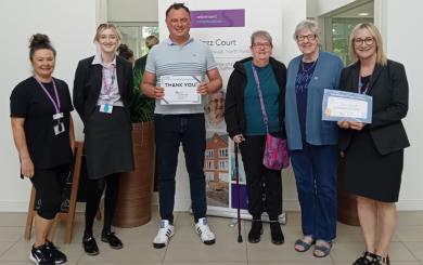Five women standing in a row with one man in the centre, smiling at the camera and holding a cheque.
