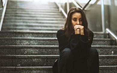 A woman in her 20s wearing dark clothes and with long dark hair, sat on the dark stairs like those which lead down to an underground station or walkway. She looks worried and has her hands clenched in front of her face.